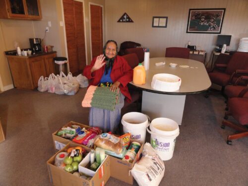 Seated elder person sits next to a table, smiling and waving to the camera. They are in a home and have three cardboard boxes with fresh produce in front of them, as well as a grain bag and two white buckets.