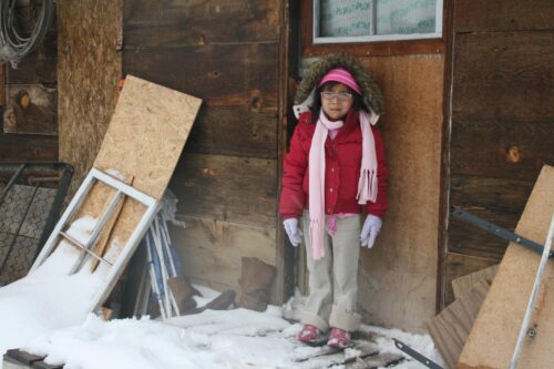 A young child stands outside a building, dressed in winter clothing, including a pink puffer jacker and a pink scarf. The ground around them is covered in snow.