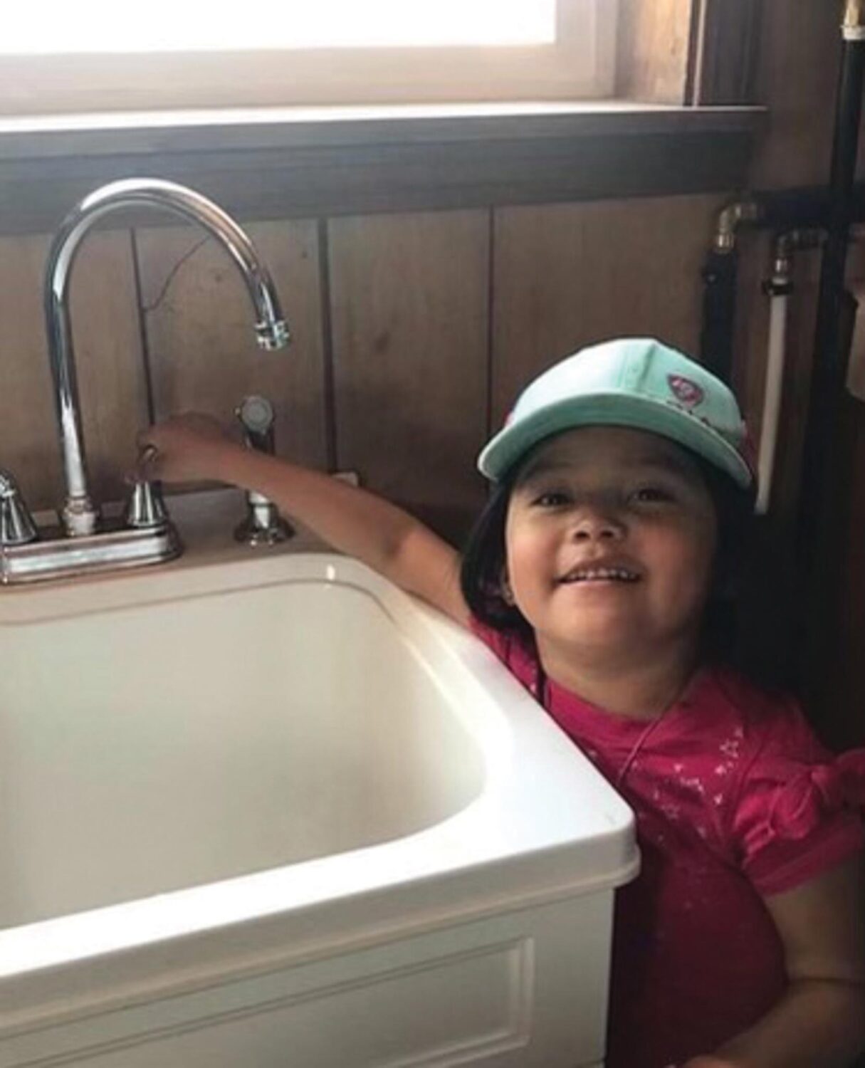 Young child with blue hat and pink shirt turns handle on for steel sink faucet