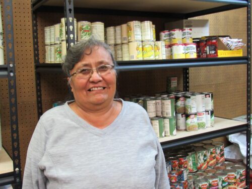 Person in grey shirt faces and smiles at the camera. Behind them, there is a black pantry shelf stocked with canned foods.