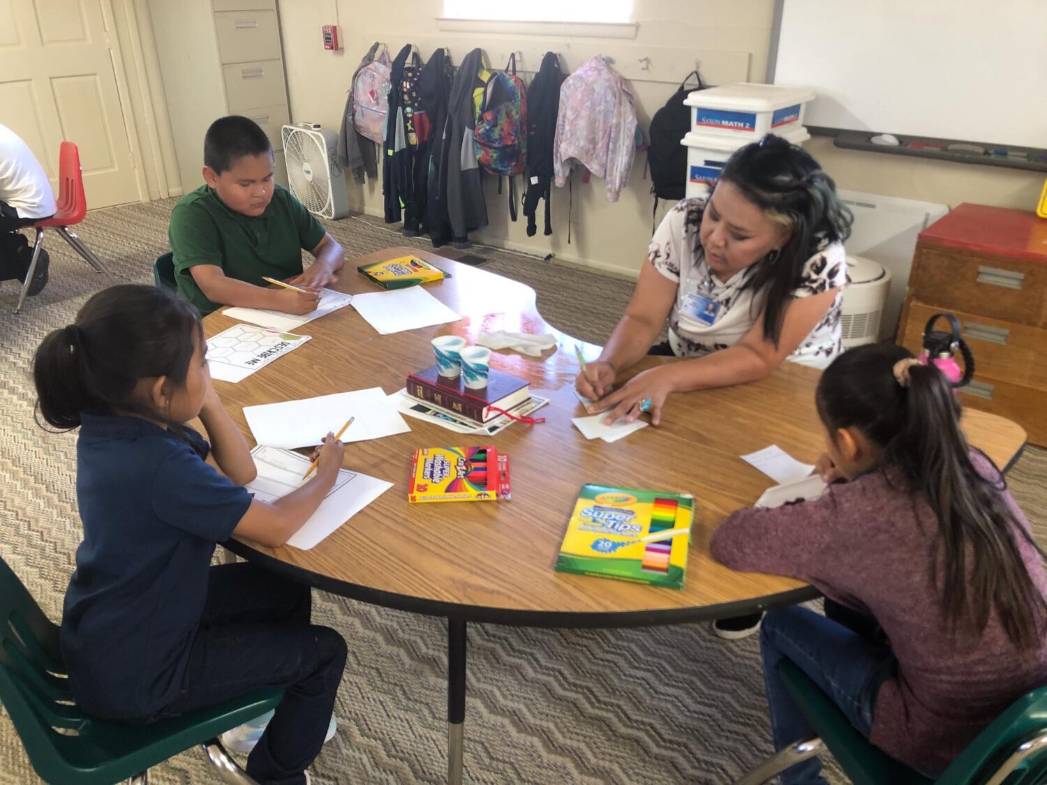 Three school-age children and one teach sit at a round classroom table, drawing with markers