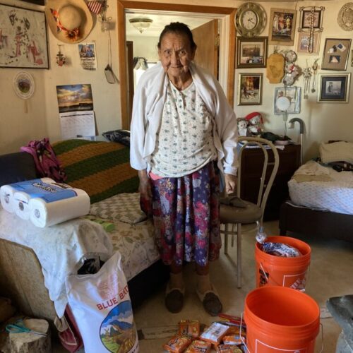 Standing elder person leans on a cane next to their bed, smiling to the camera. They are in a home and have a new roll of paper towels, a grain bag, and 2 orange buckets next to them.