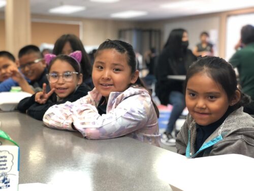 A group of smiling children sit at a cafeteria table, with three of them in the foreground looking toward the camera. One child wears cat-ear headbands and glasses, and others wear colorful jackets.