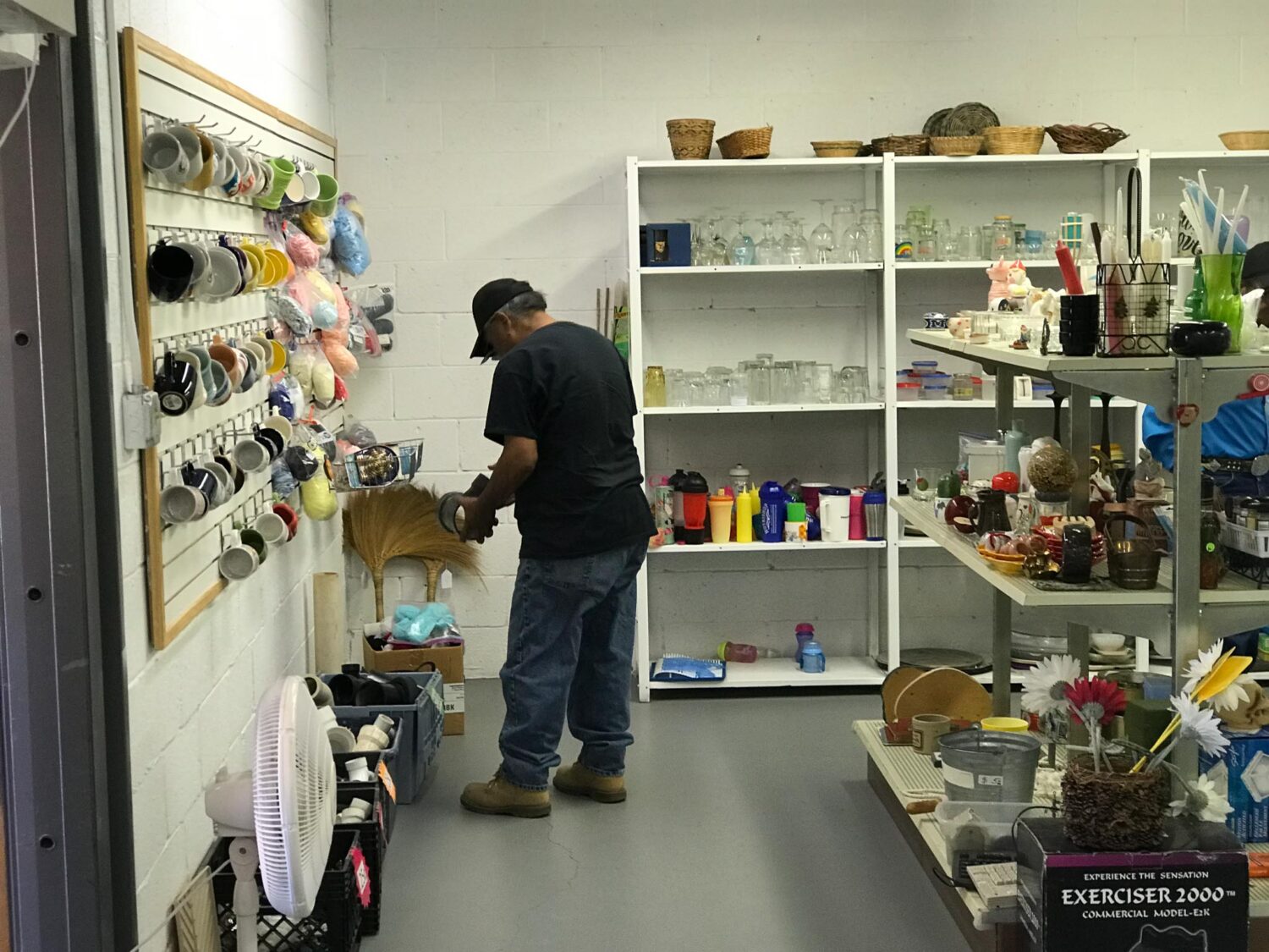 A person wearing a baseball cap is holding and inspecting an item in the corner of a stocked pantry. The pantry includes a range of household items, including mugs and glassware.