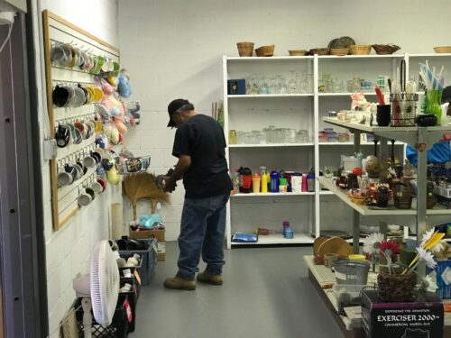 A person wearing a baseball cap is holding and inspecting an item in the corner of a stocked pantry. The pantry includes a range of household items, including mugs and glassware.