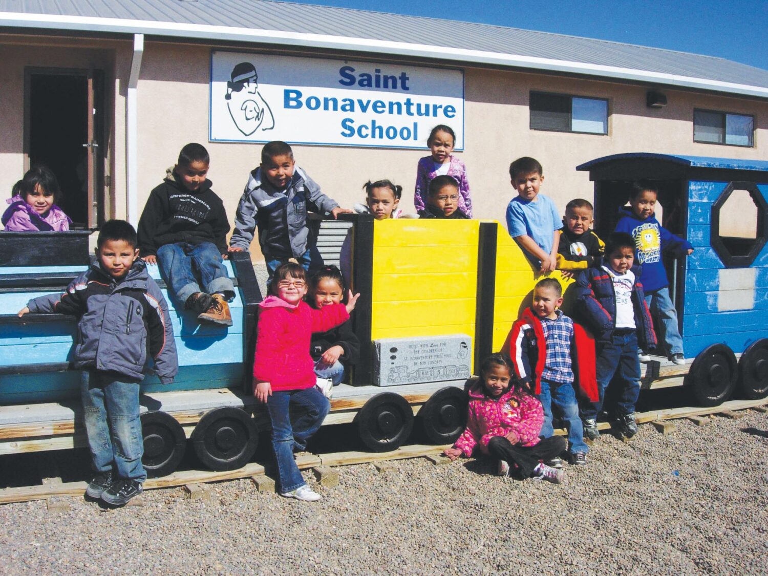 A group of young children smile towards the camera, posed along the length of blue child-sized toy train. Some are seated in front of or on top of the train while others lean against it. The train is parked in front a building with a white sign that says 