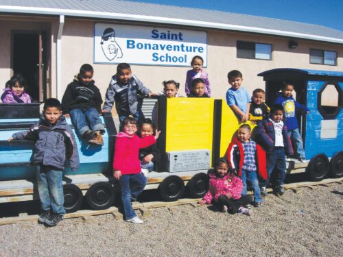 A group of young children smile towards the camera, posed along the length of blue child-sized toy train. Some are seated in front of or on top of the train while others lean against it. The train is parked in front a building with a white sign that says 