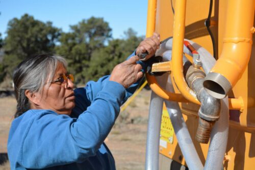 A person in a blue sweatshirt connects a hose to a large yellow water delivery truck under a clear blue sky, surrounded by trees.