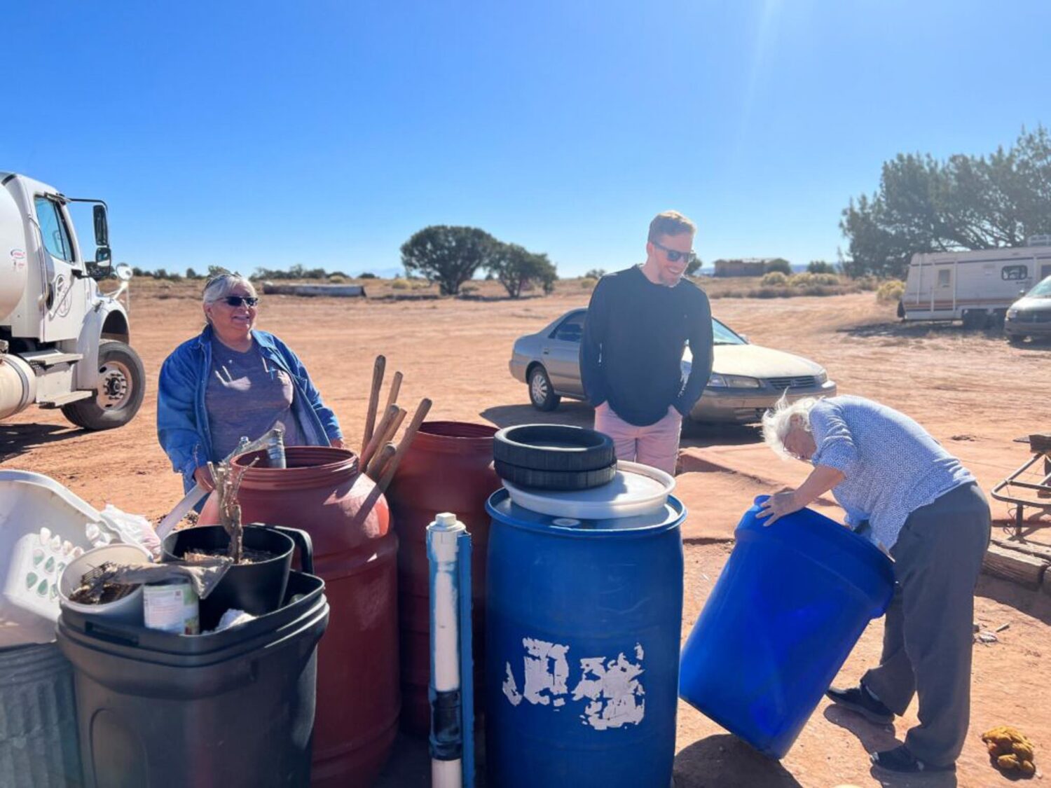 Three people stand outside under a sunny sky, surrounded by plastic water tanks. One person tips and peeks into a blue water tank. The front of water delivery truck is seen behind them.