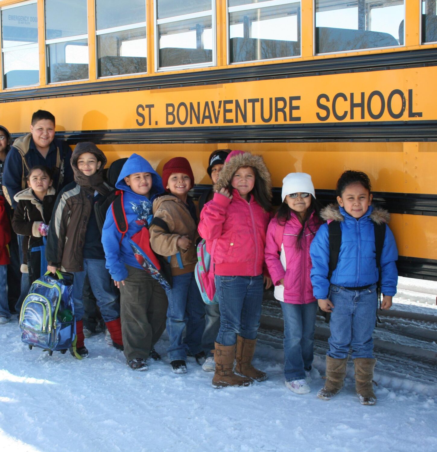Group of young school-age children stand outside in their winter clothes in front of a yellow school bus with the title